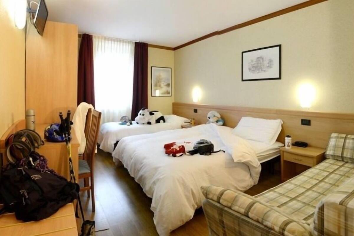 A hotel room with two beds, a wooden wardrobe, a desk with a chair, and a small table with a lamp.