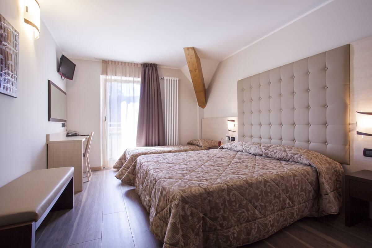 A bedroom with two beds, a desk, a bench, and a TV on the wall.