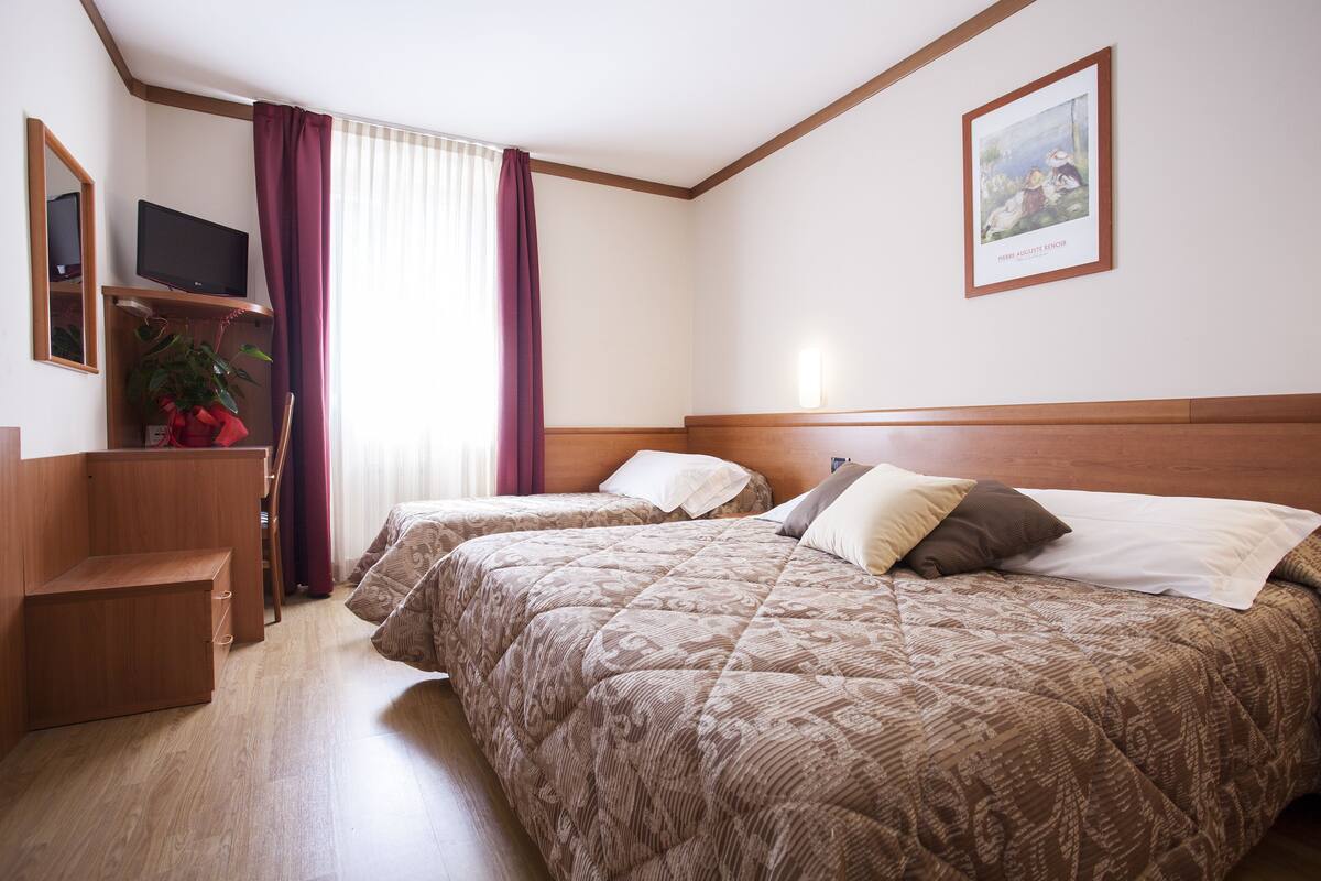 A hotel room with two beds, a TV on a shelf, a desk, and a window with curtains.