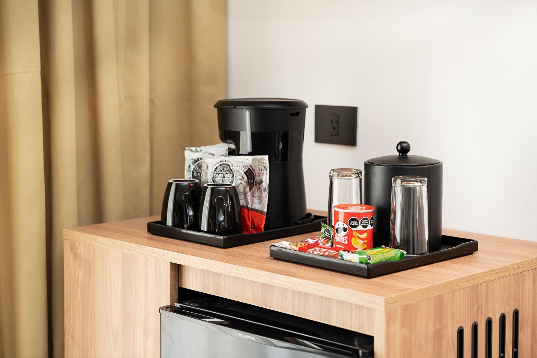 A coffee station with a coffee maker, mugs, a thermos, and snacks on a wooden counter.