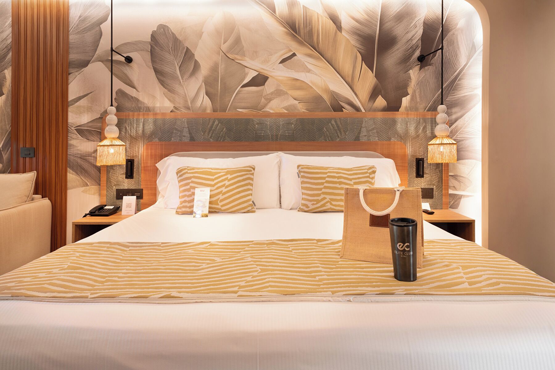 Habitación de hotel con una cama grande, dos lámparas de noche, una mesita de noche, una pared con un diseño de hojas y una maleta sobre la cama.