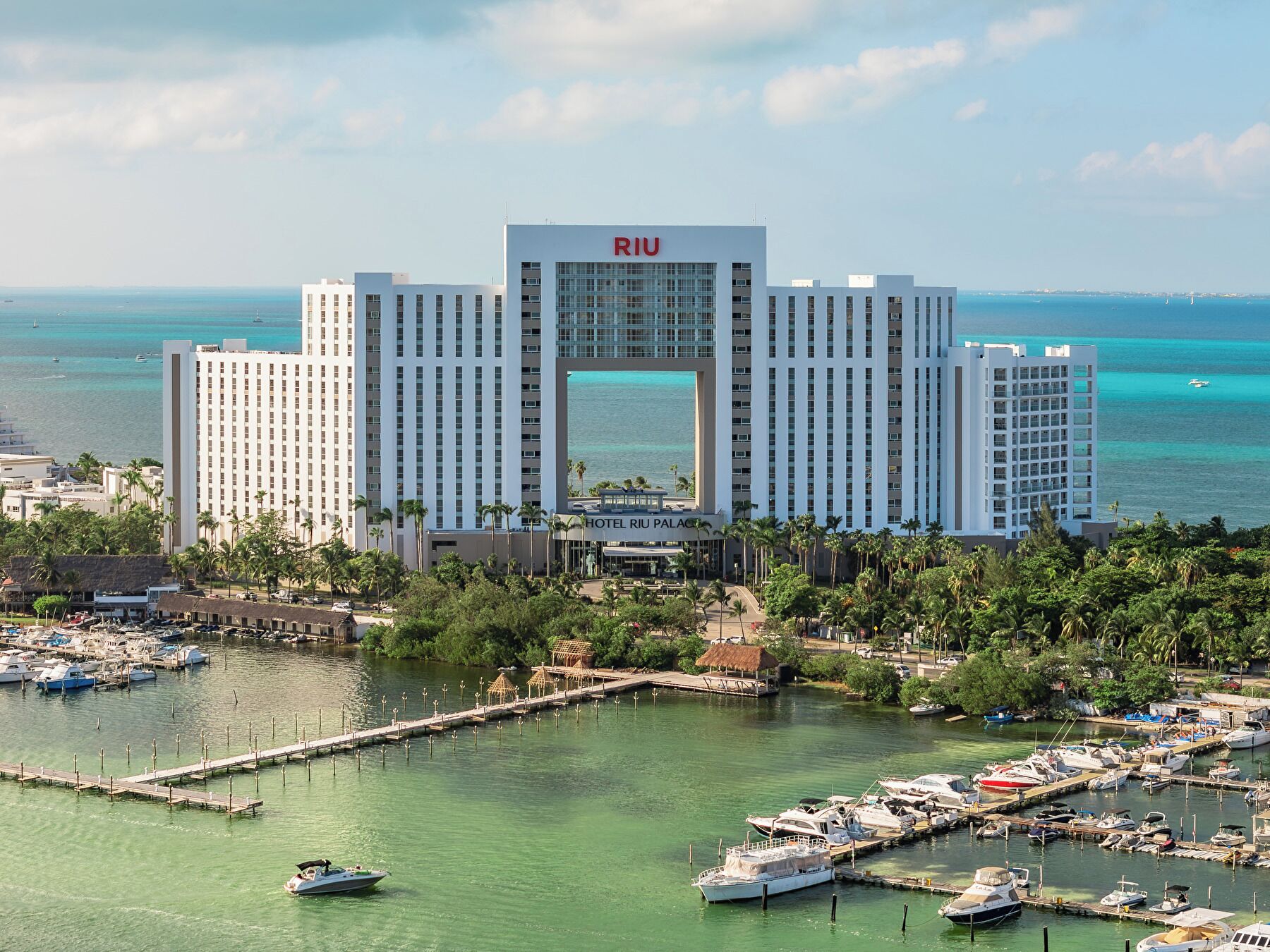 A large hotel with a prominent central entrance, surrounded by a marina with boats and a clear view of the ocean.
