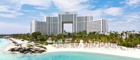 A large hotel with a central archway, surrounded by a beach and palm trees.