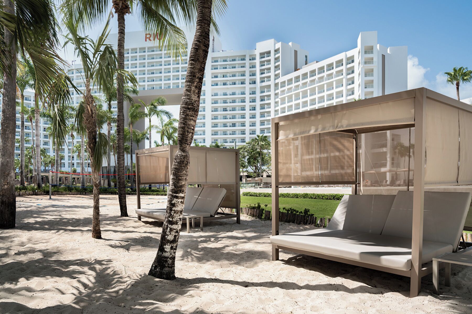 Una zona de playa con palmeras, arena y una hilera de edificios blancos al fondo.