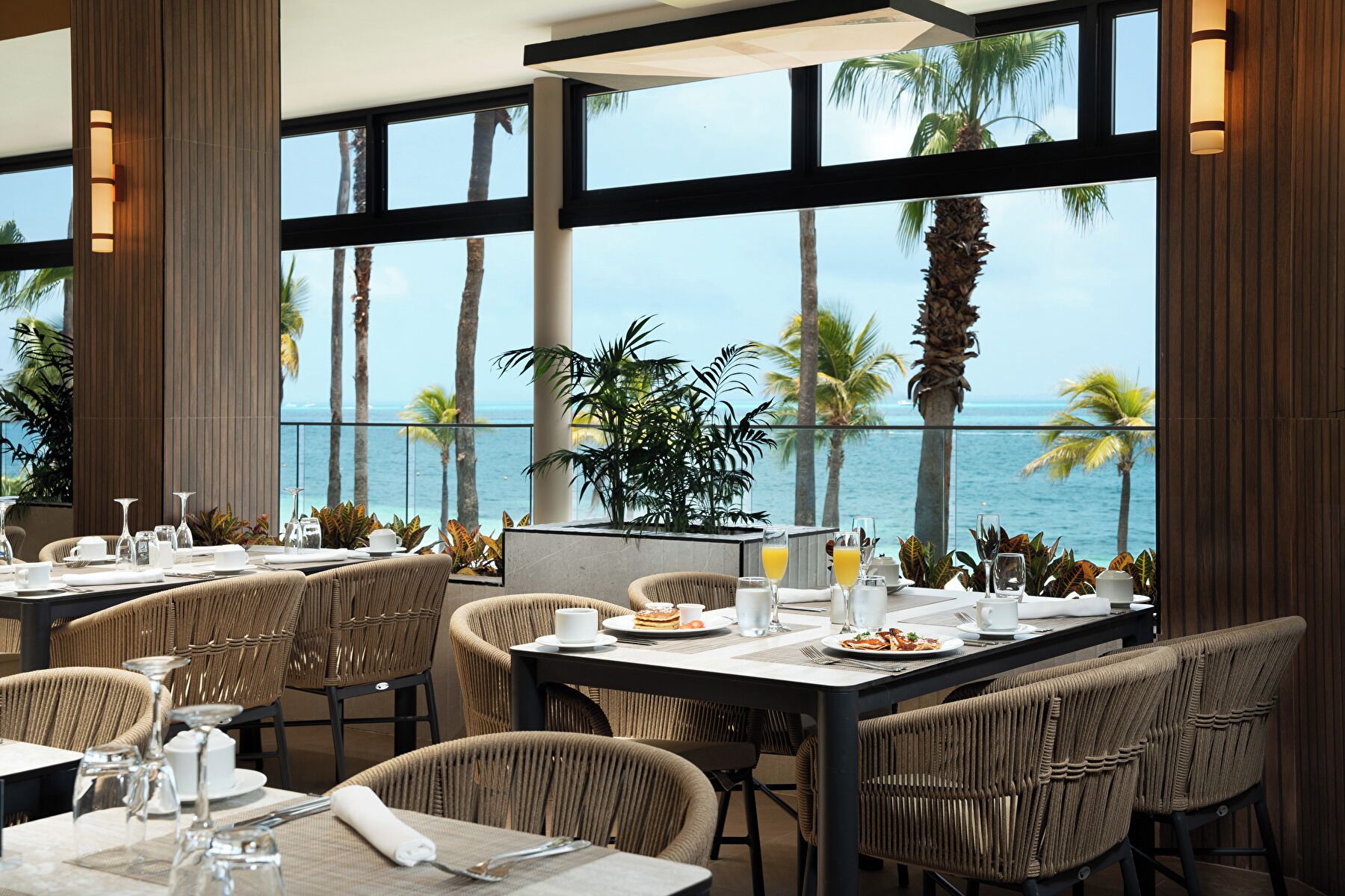Comedor con mesas dispuestas para dos, con vista a una playa con palmeras y el océano.