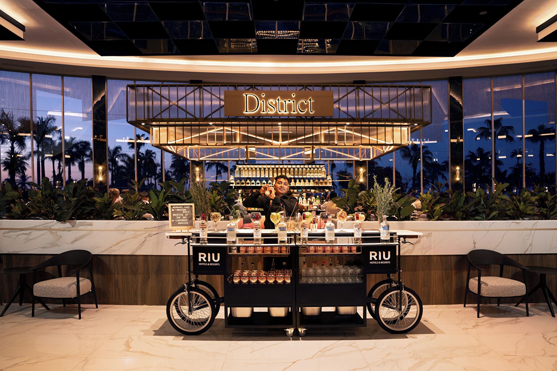 A bar area with a sign reading 'District' and a cart labeled 'RIU Hotels & Resorts' filled with bottles and glasses.