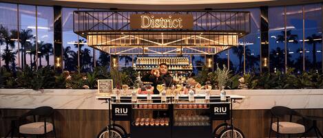 A bar area with a sign reading 'District' and a cart labeled 'RIU Hotels & Resorts' filled with bottles and glasses.