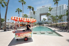 A poolside area with a food cart labeled 'Bionicos,' palm trees, and a hotel building with 'RIU' signage. - Hotel Riu Palace Peninsula - All Inclusive (Cancun)