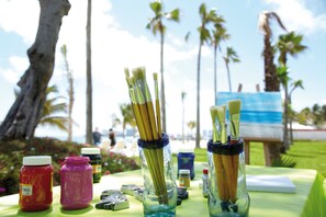 A table with paint jars and brushes, a painting in progress, and a tropical outdoor setting. - Hotel Riu Palace Peninsula - All Inclusive (Cancun)