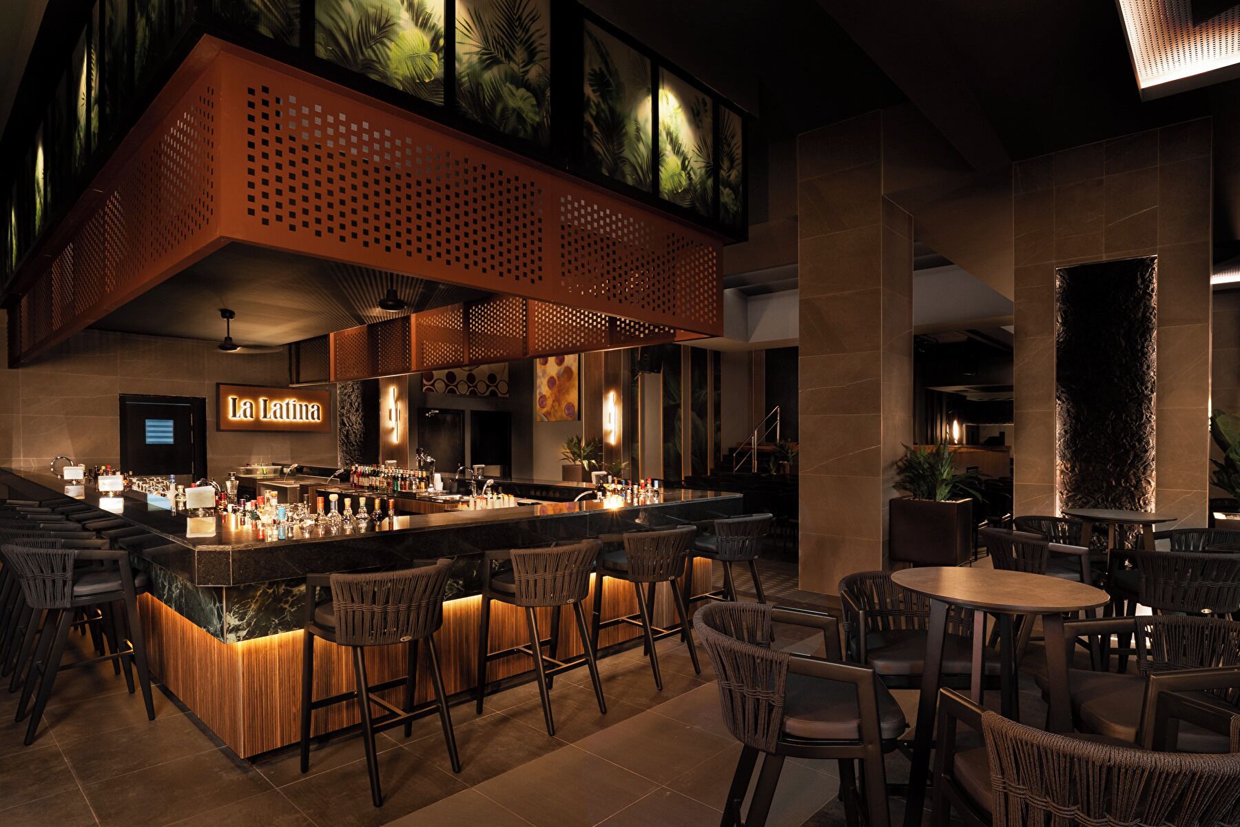 Zona de bar con techo alto, una barra con diferentes botellas y disposición de mesas y sillas.