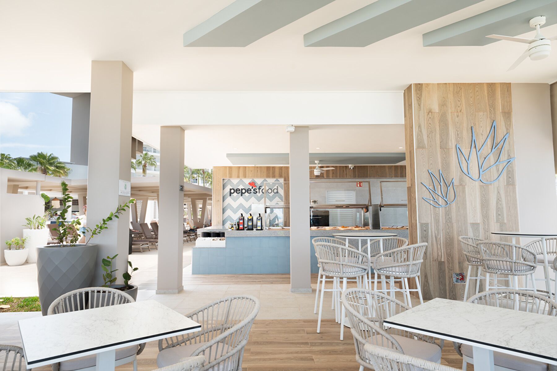 Un área de comedor moderna al aire libre con sillas de mimbre y mesas blancas, una zona de bar con un letrero 'pepe'sfood', y vista al cielo.