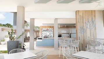 Un área de comedor moderna al aire libre con sillas de mimbre y mesas blancas, una zona de bar con un letrero 'pepe'sfood', y vista al cielo.