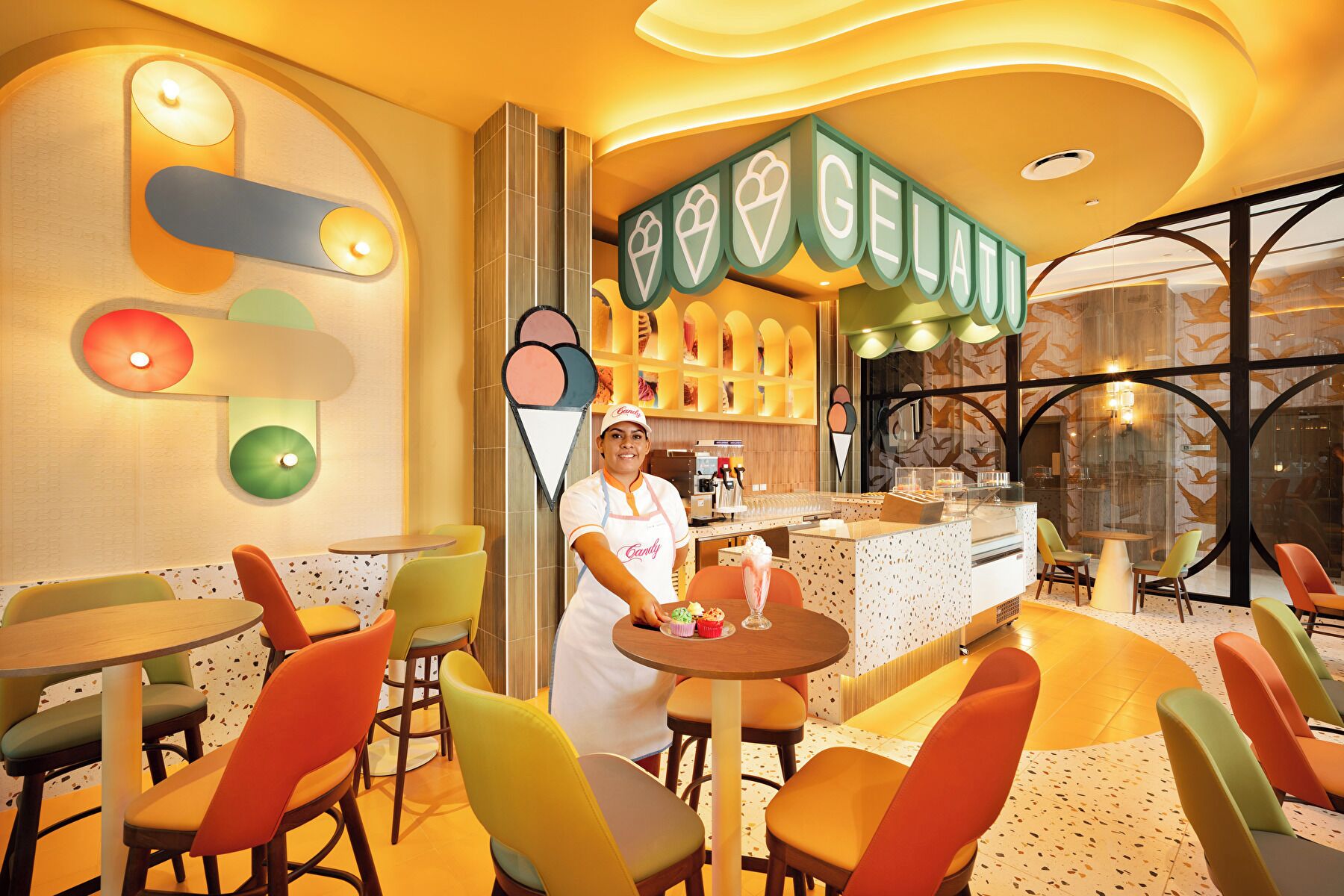 Une pâtisserie lumineuse, avec des chaises et des tables aux couleurs vives, un comptoir où sont présentés des cornets de glace, et une personne qui sert les desserts.