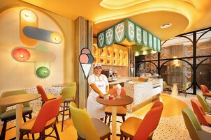 Una pastelería luminosa con sillas y mesas coloridas, una barra con helados expuestos y una persona atendiendo.