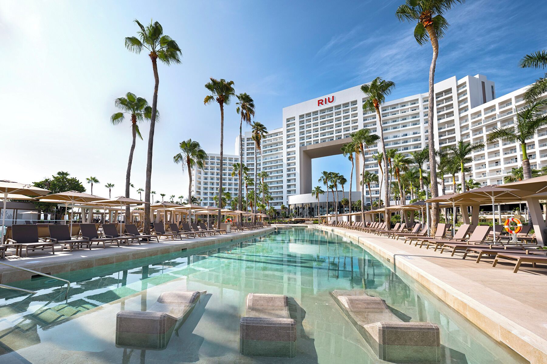 A hotel with a large pool, palm trees, and a clear sky.