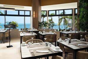 Un restaurante con mesas dispuestas para comer, amplias ventanas que ofrecen vista al océano y un cartel que indica una opción especial de comida.