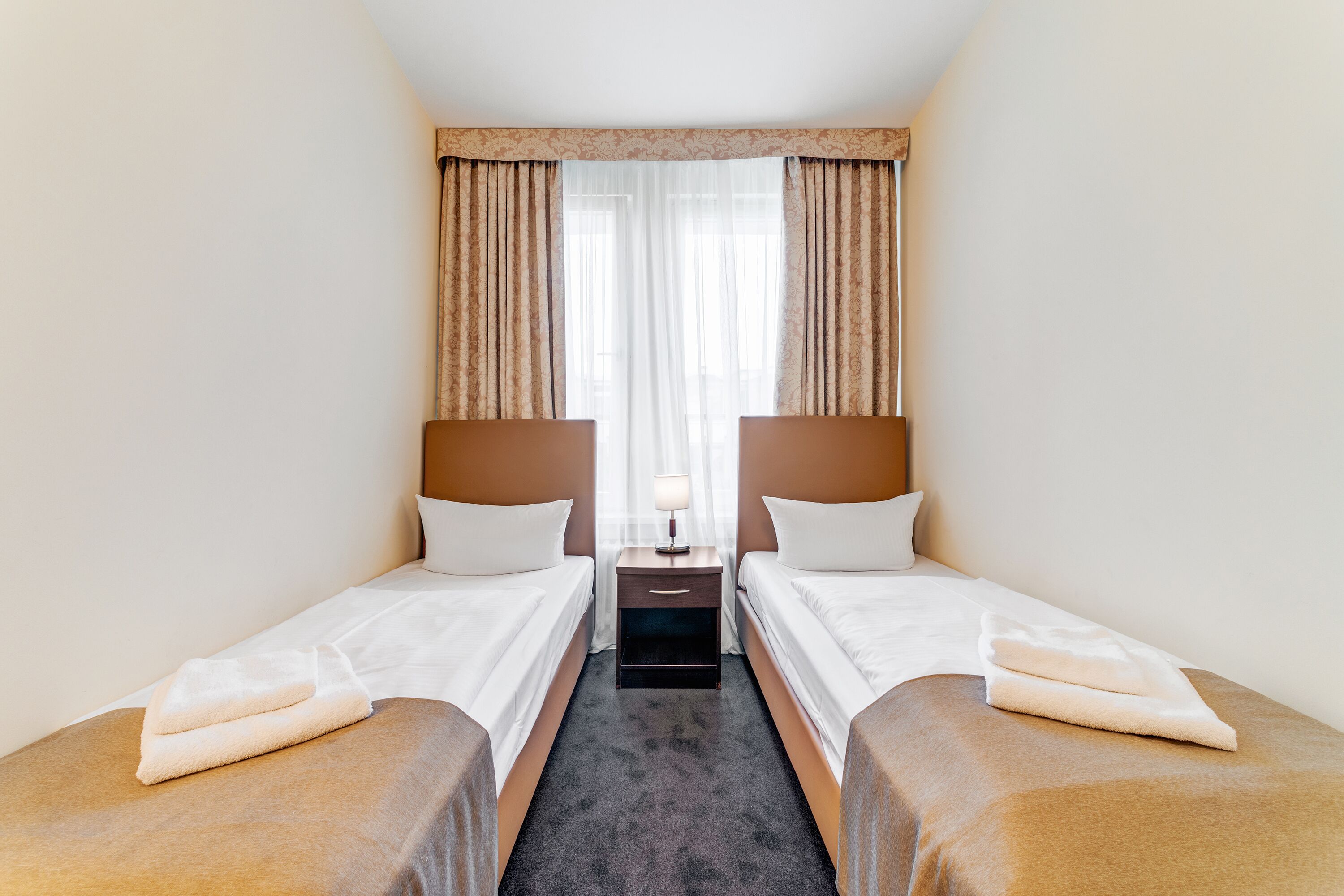 Een hotelkamer met twee enkele bedden, een nachtkastje met een lamp en een raam met dunne gordijnen.