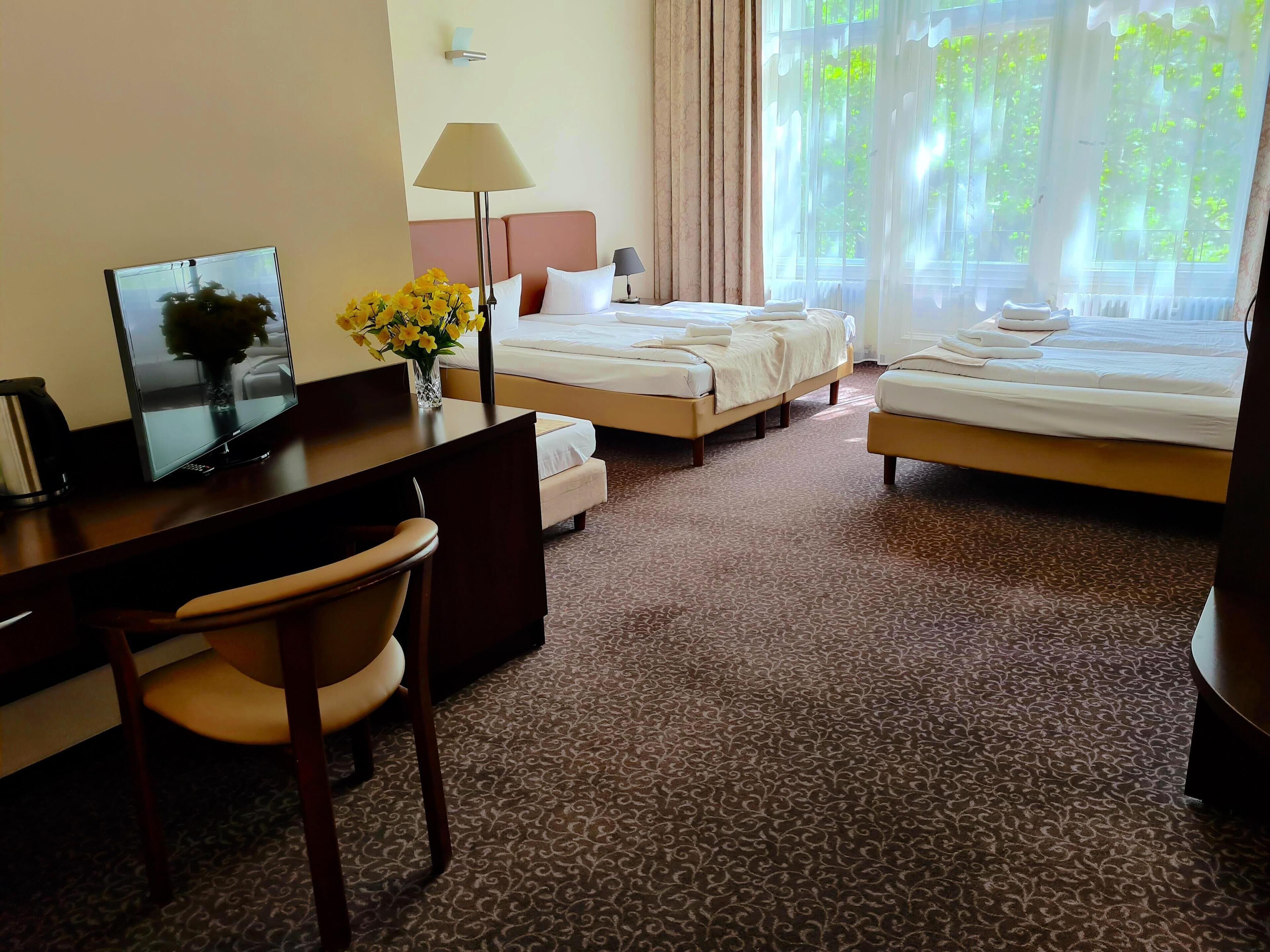 Habitación de hotel con dos camas, un escritorio con silla, televisión, lámpara y un florero con flores.