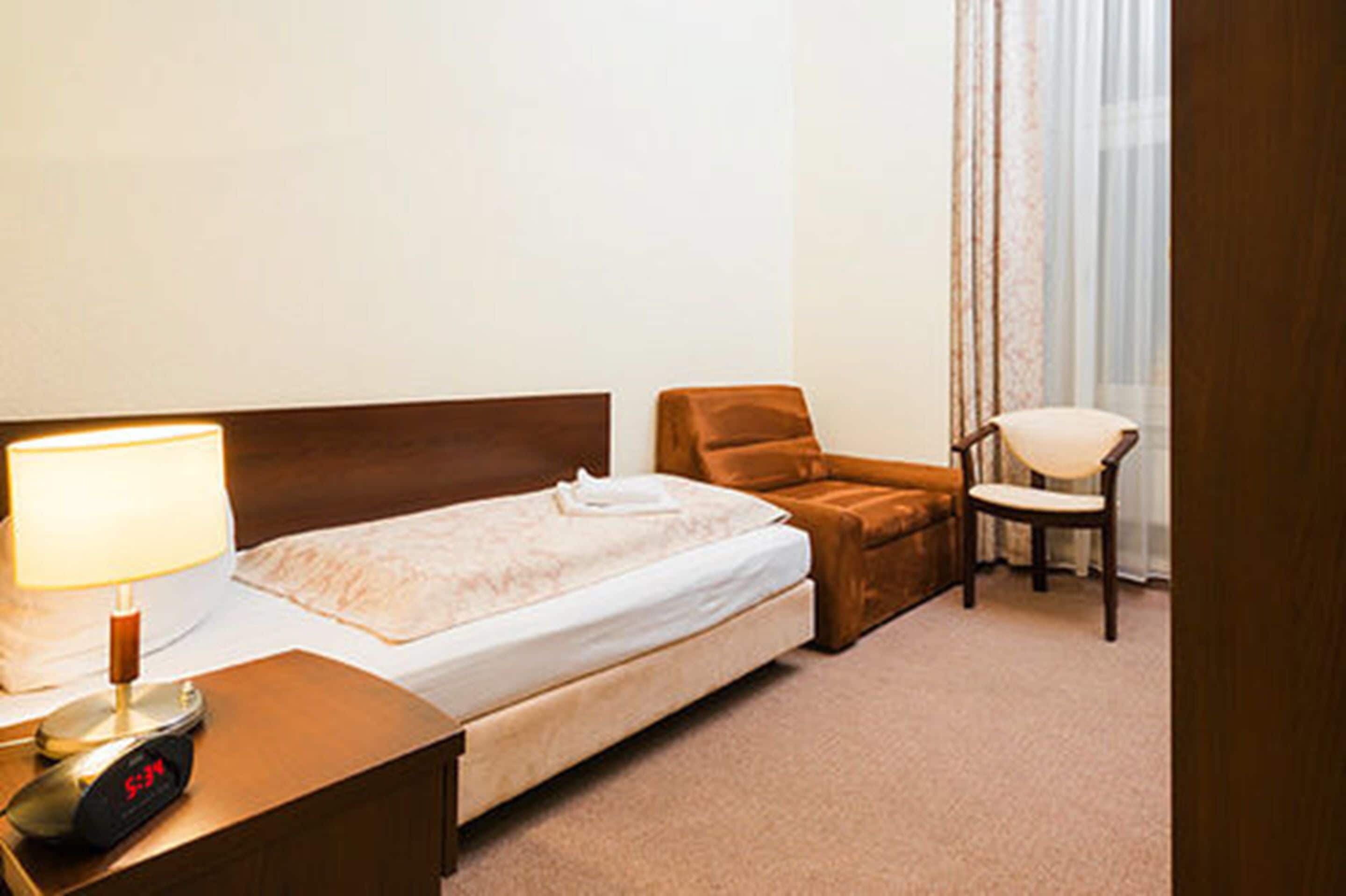 Una habitación de hotel con cama, una silla, un escritorio con lámpara y un reloj.
