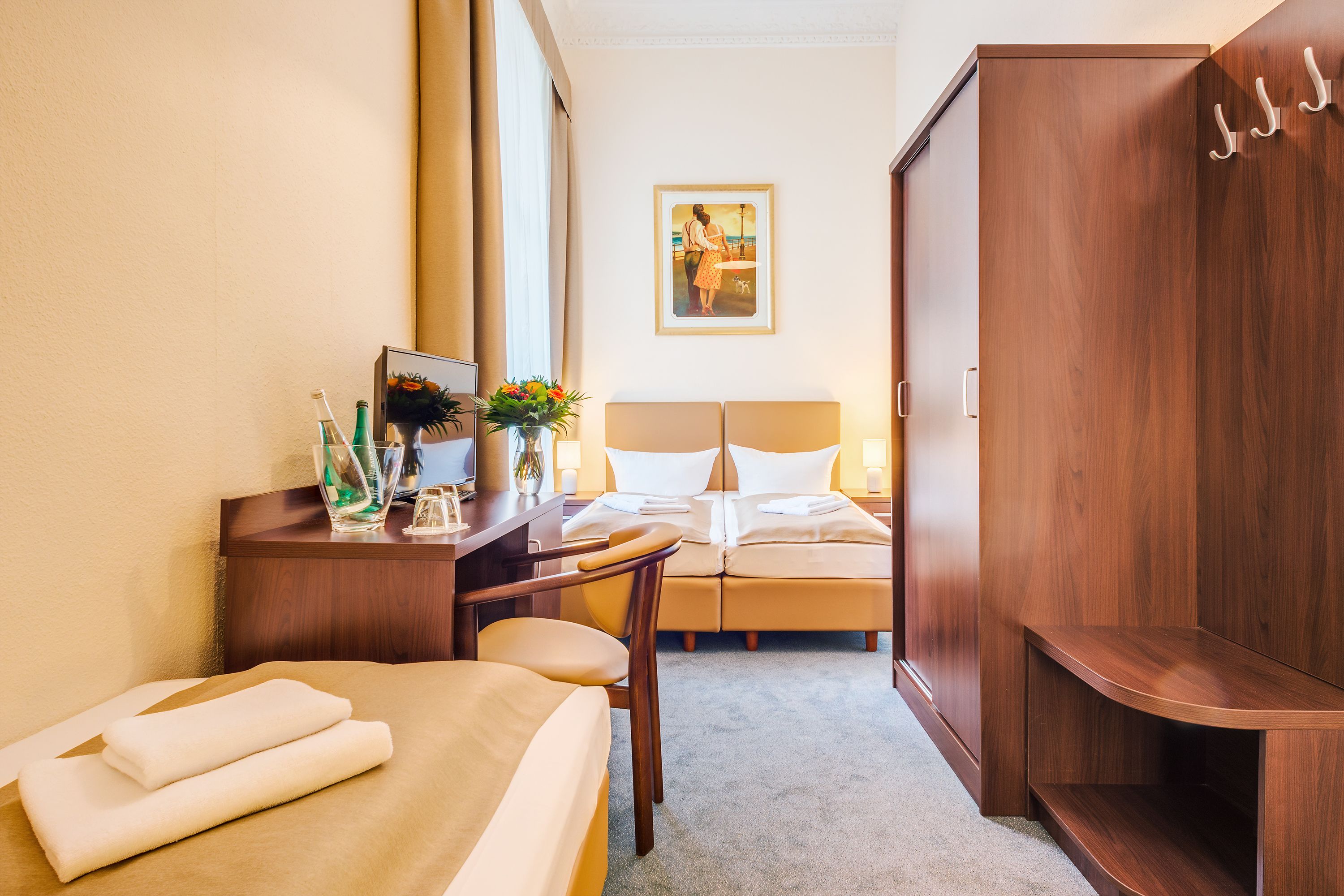 Une chambre d’hôtel avec un lit, un bureau agrémenté d’un vase de fleurs, un cadre photo et une armoire en bois.