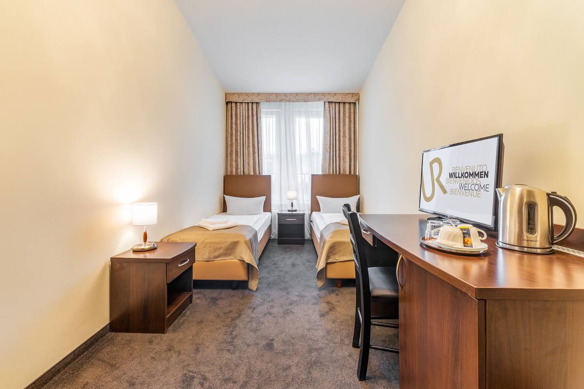 Una camera d'albergo con due letti, una scrivania, una sedia, una macchina per caffè e un cartello di benvenuto.
