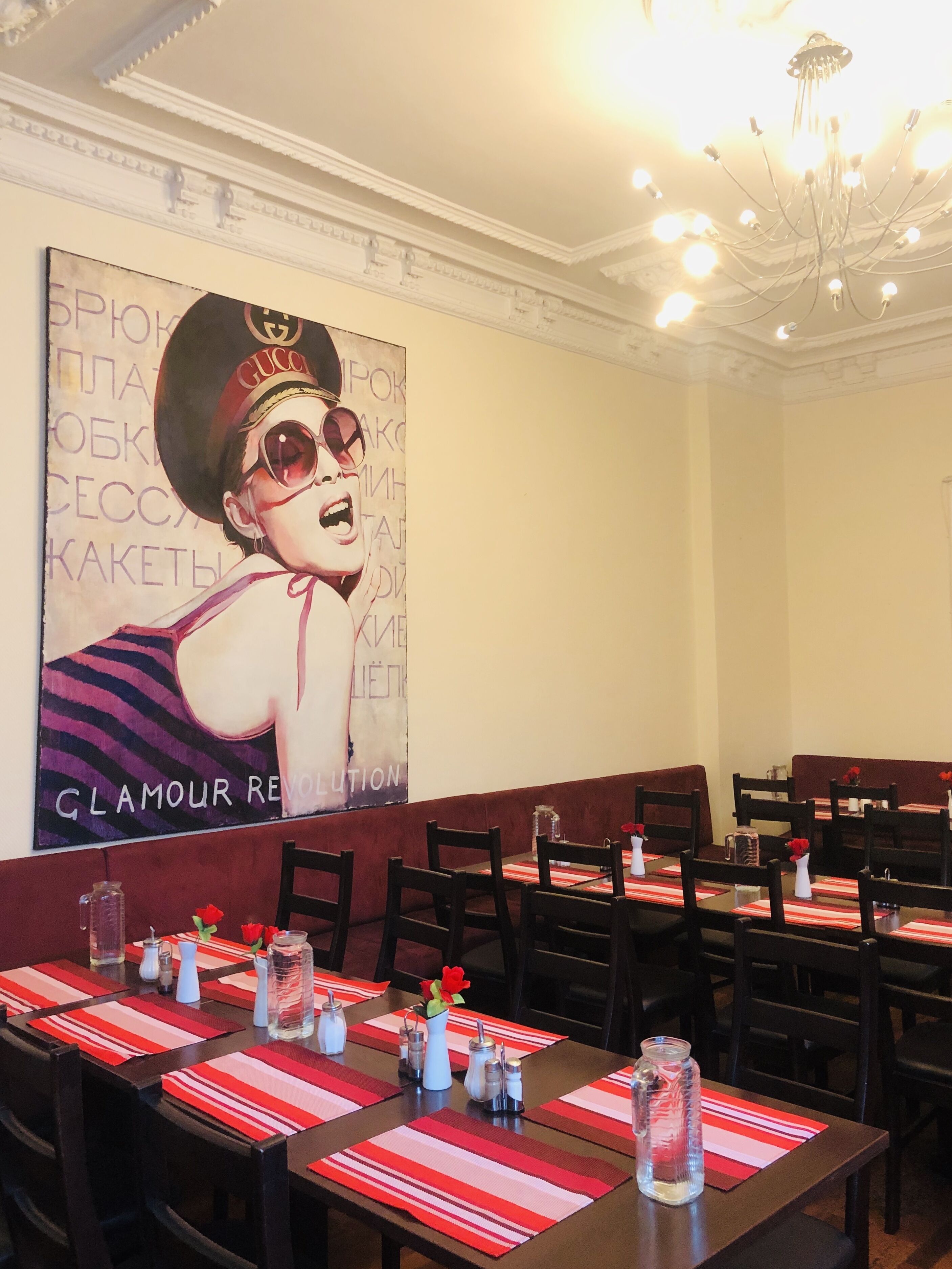Un restaurant où les tables sont dressées avec des sets de table rayés rouge et blanc, des roses rouges et des carafes en verre transparent. Une grande fresque murale représente une personne portant un chapeau et des lunettes de soleil de la marque Gucci.