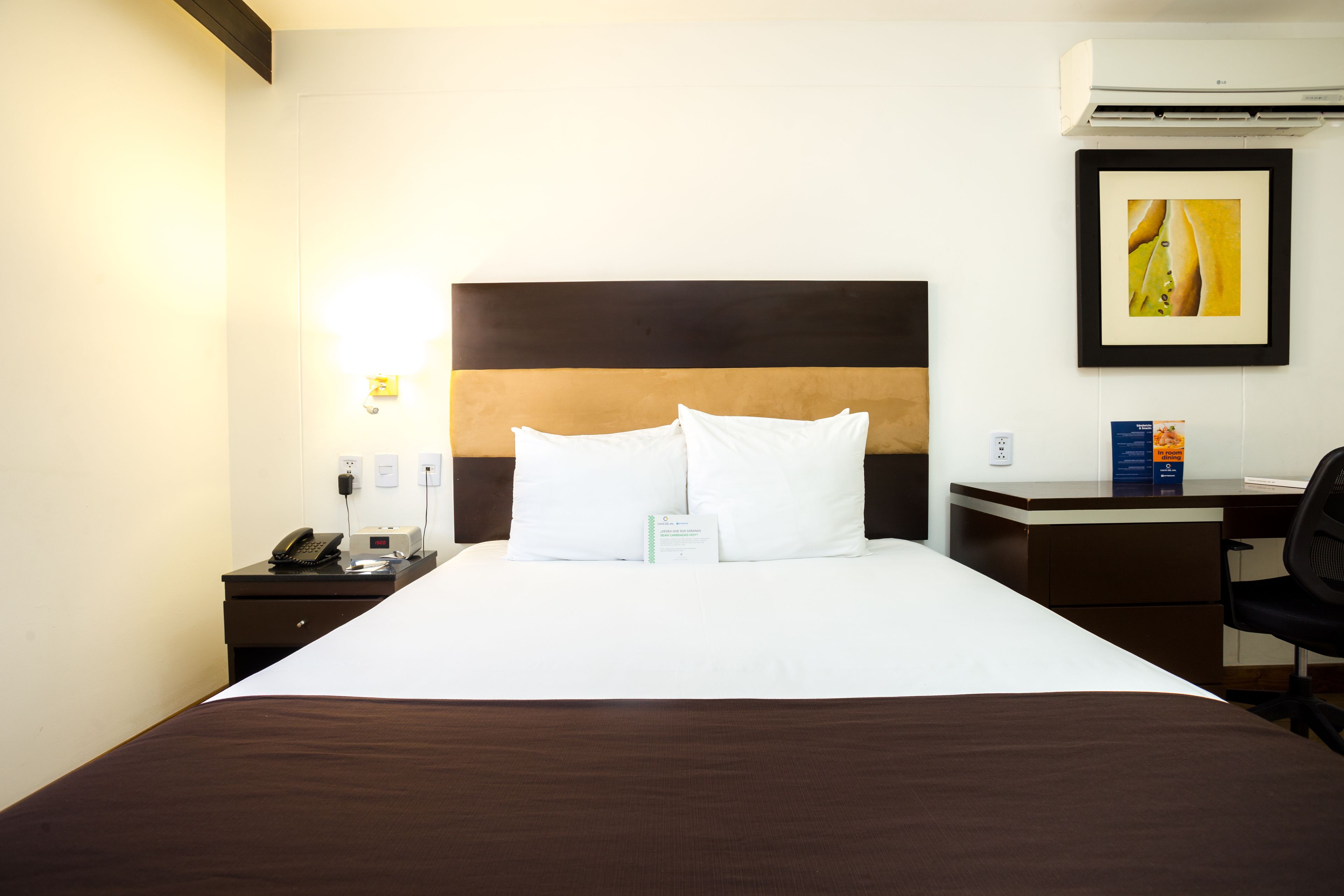A hotel room with a large bed, a desk, a chair, a phone, and a framed picture on the wall.