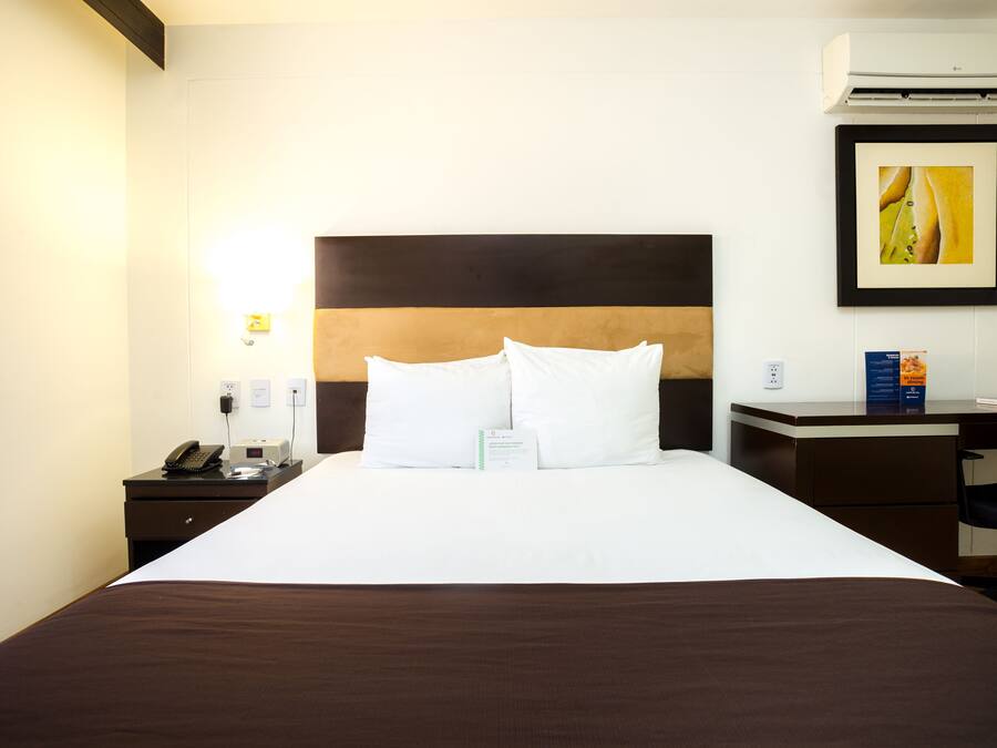 A hotel room with a large bed, a desk, a chair, a phone, and a framed picture on the wall.