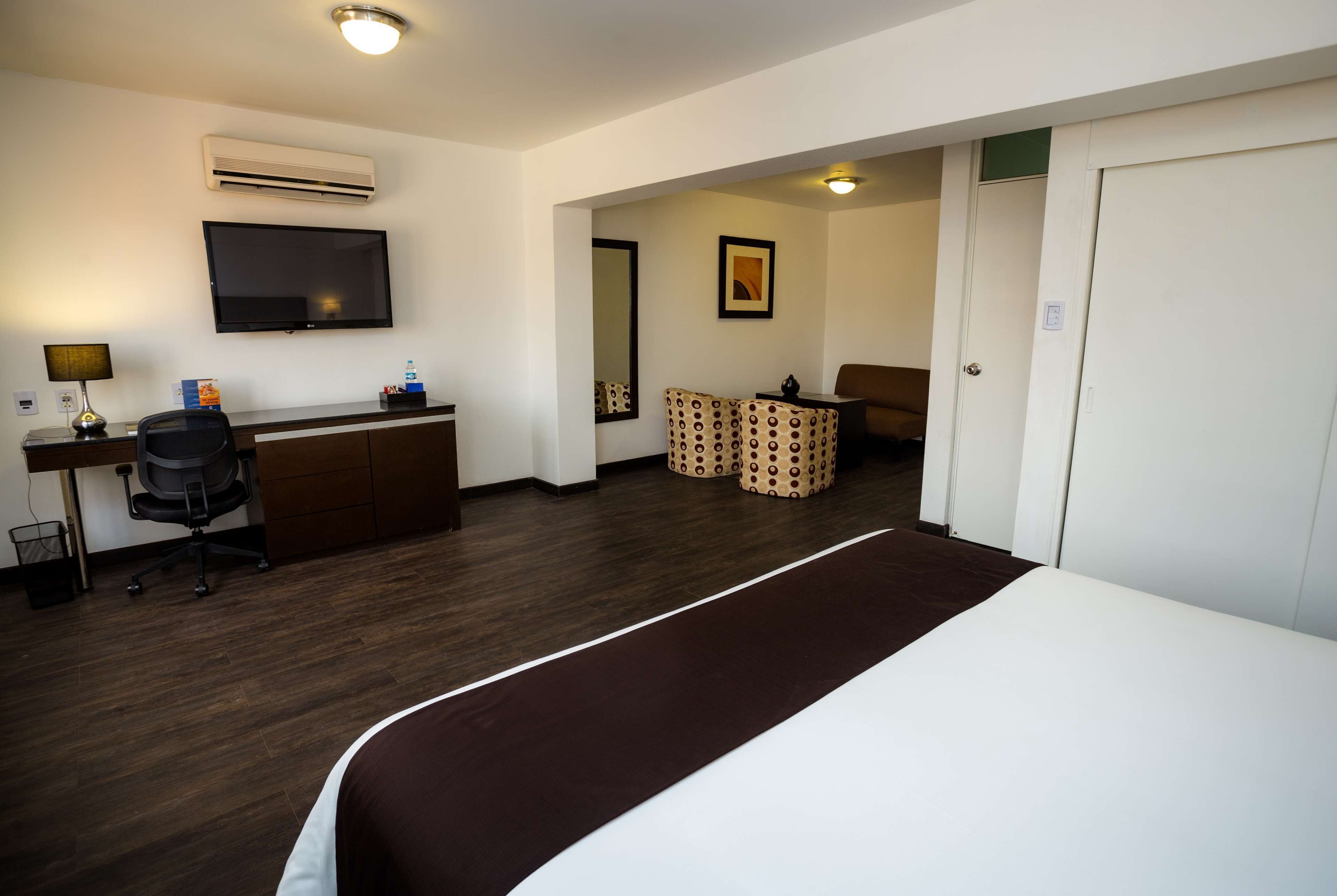 A hotel room with a bed, desk, chair, TV, air conditioning unit, and a door.