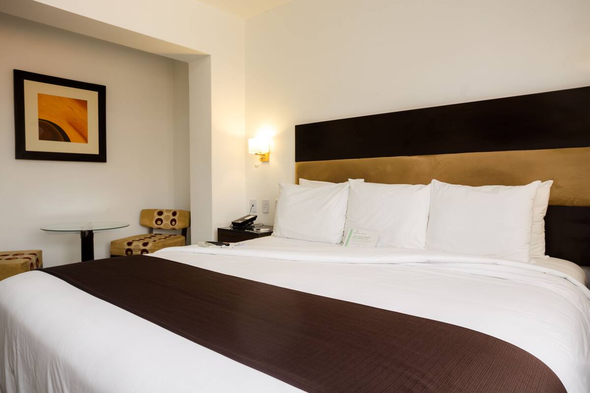 A hotel room with a large bed, a bedside table, a chair, and a wall-mounted light.