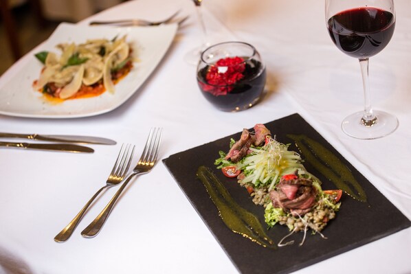 Una mesa de comedor con un plato de pasta, una copa de vino tinto y un plato negro con un guiso de carne.