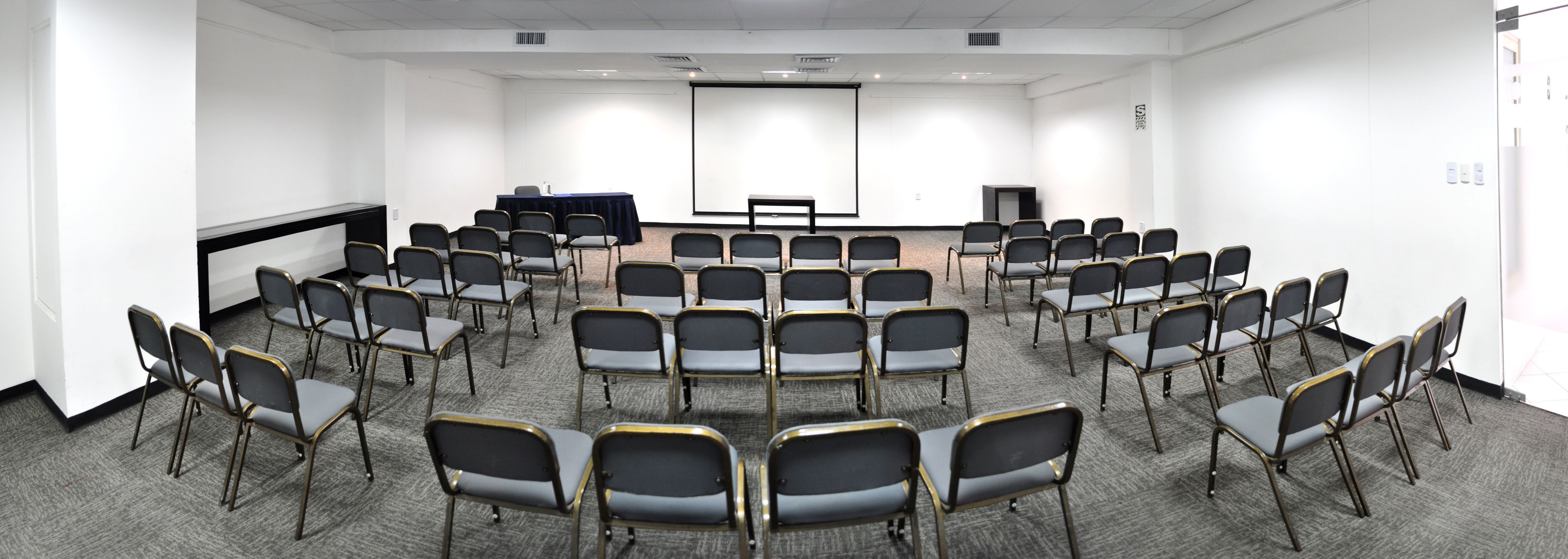 A conference room with rows of chairs facing a projector screen, a table with a laptop, and a carpeted floor.