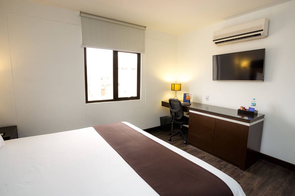 A hotel room with a bed, desk, chair, TV, and air conditioning unit.