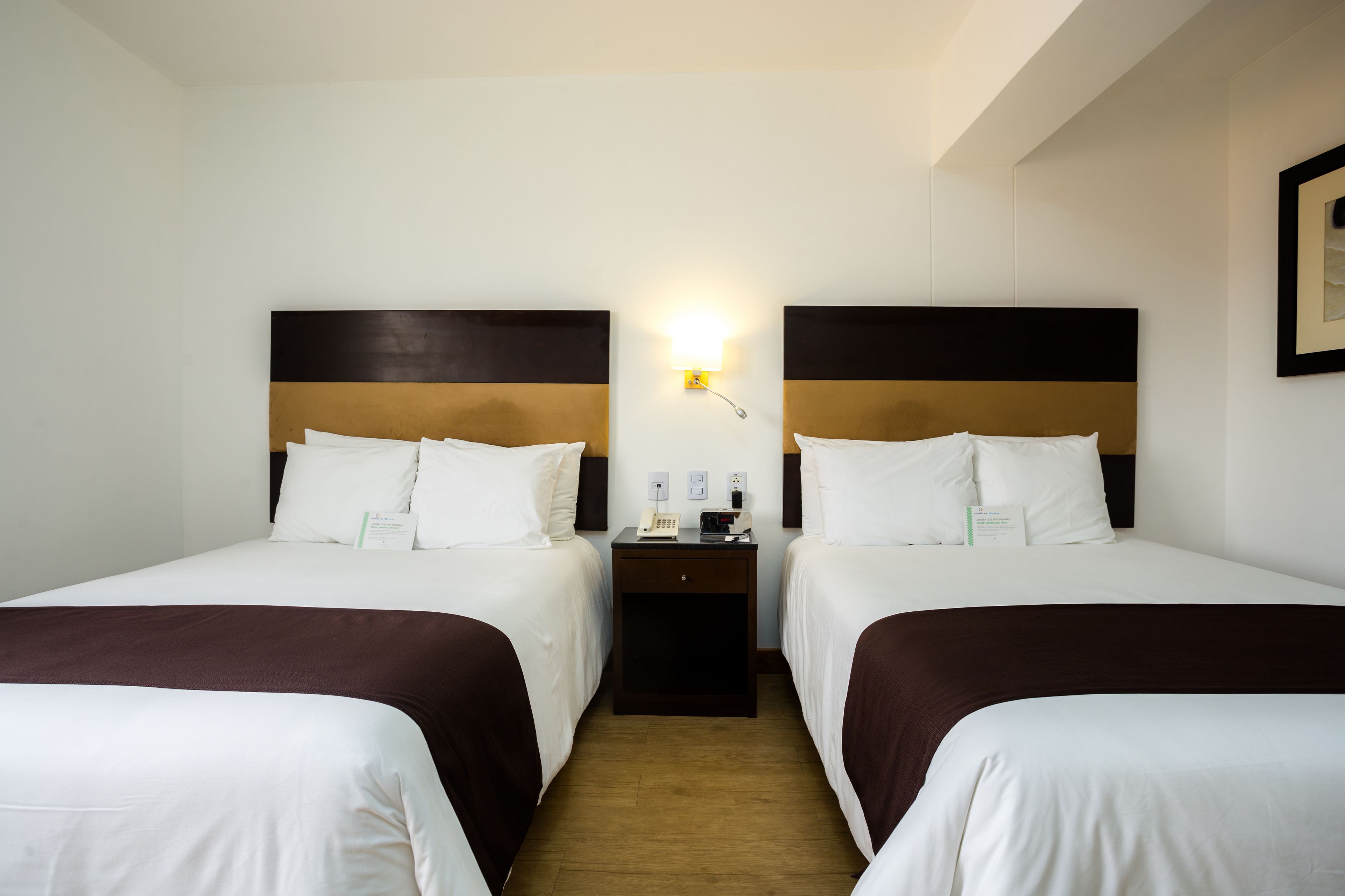 A hotel room with two beds, each with white linens and brown headboards. There is a nightstand between the beds with a phone and a small table.