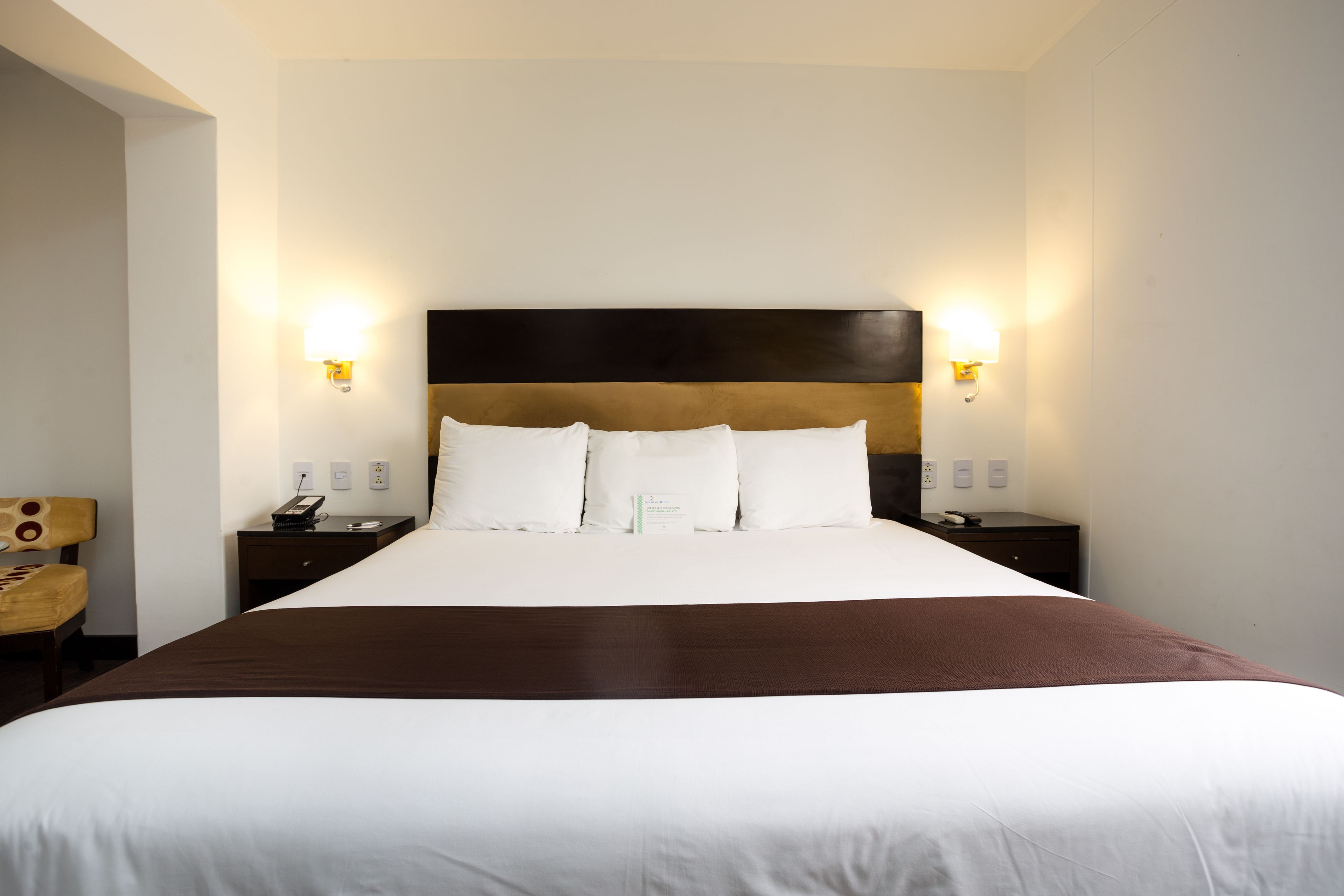 Habitación de hotel con una cama grande, dos mesitas de noche, una silla y lámparas fijadas a la pared.