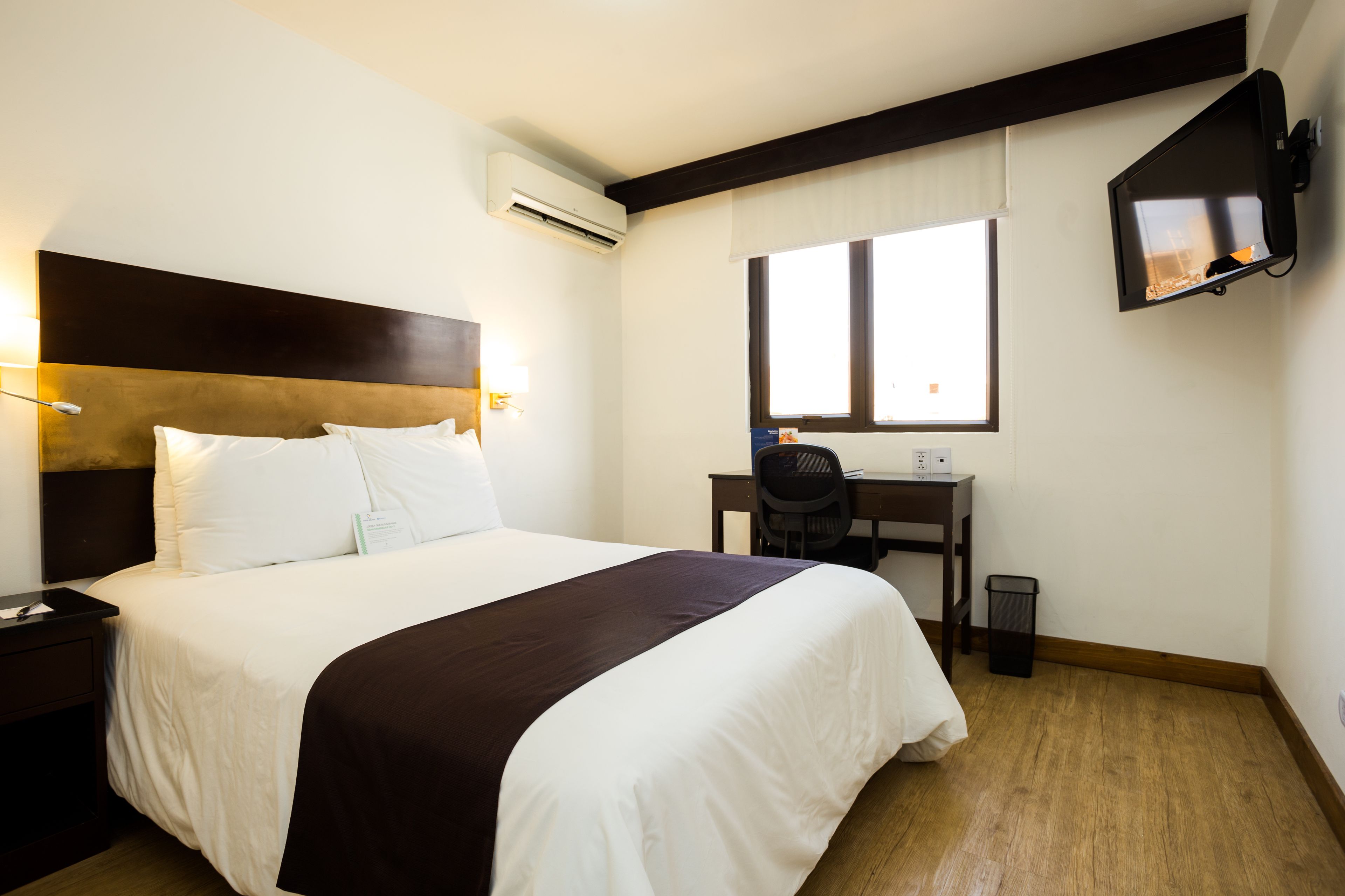 A hotel room with a large bed, a desk, a chair, and a TV mounted on the wall.