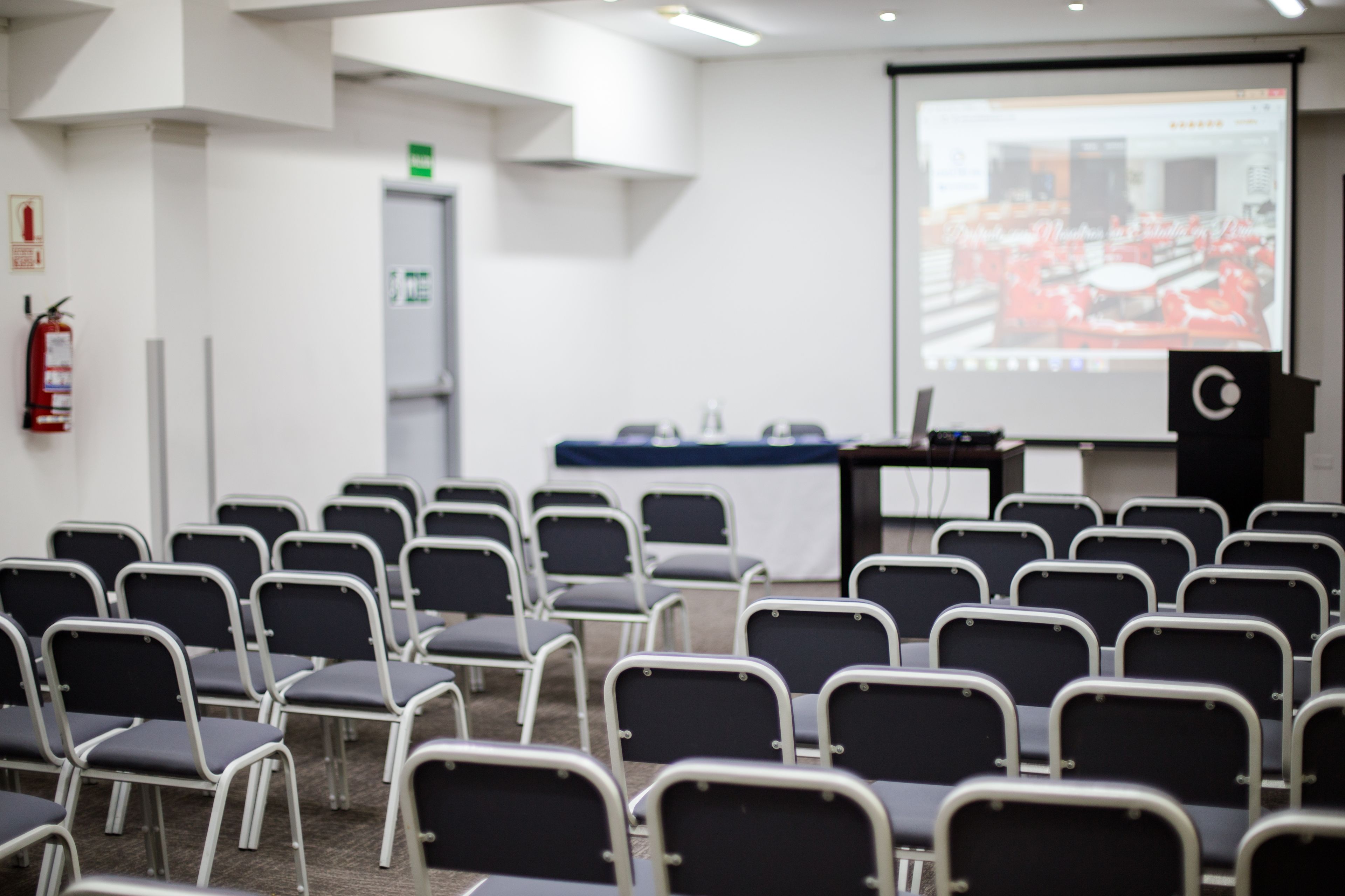 Un salón de conferencias con filas de sillas, una pantalla de proyector, un atril y un extintor.
