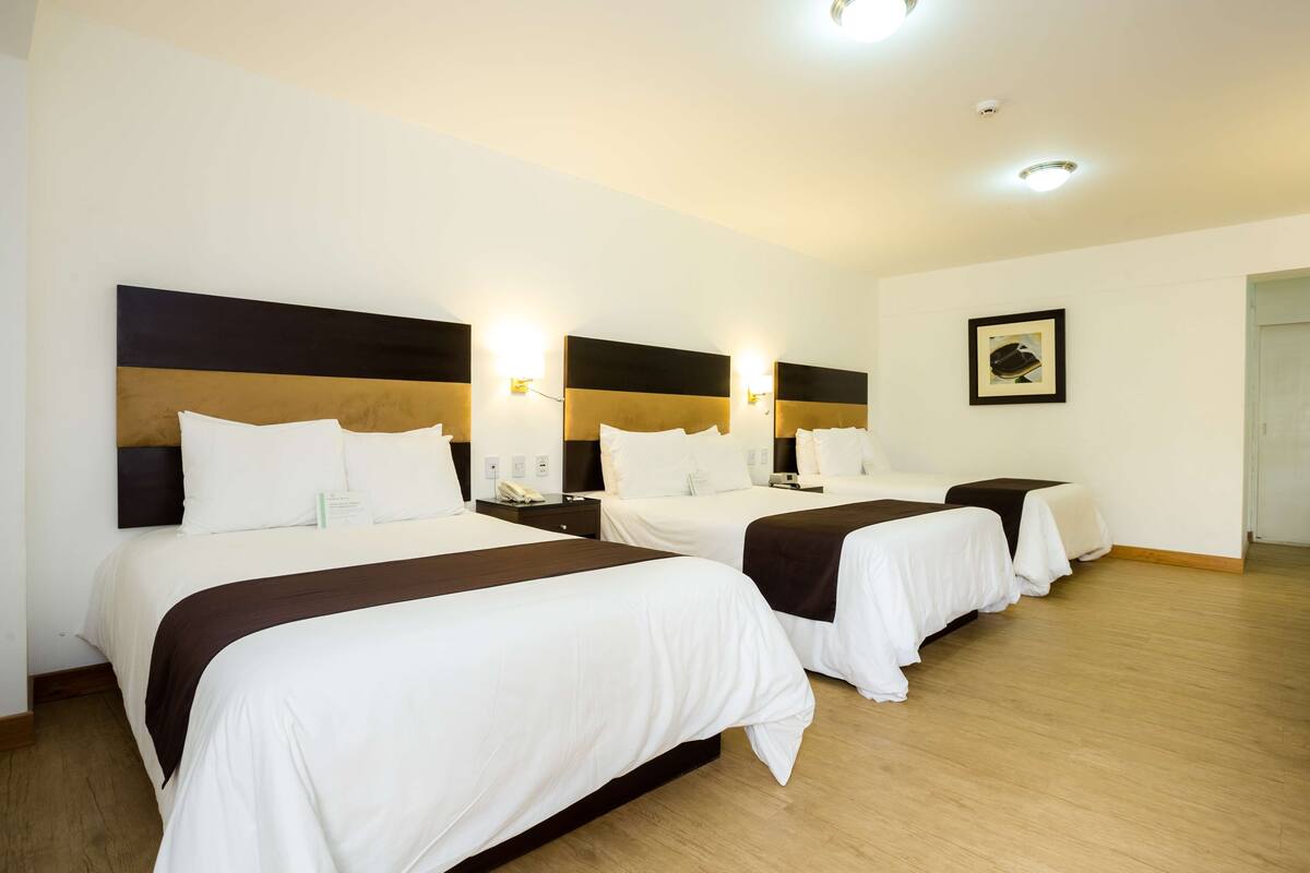 A hotel room with three single beds, each with a headboard, bedside tables, and wall-mounted lamps.