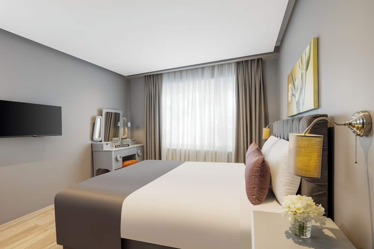 Quarto de hotel moderno com uma cama grande, uma televisão de tela plana, uma mesa de cabeceira com luminária e uma janela com cortinas.