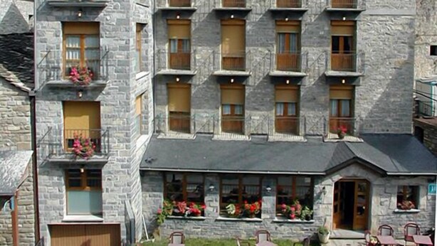 A stone building with multiple windows and balconies, a tower, and a small garden with outdoor seating. - Hotel Gabarre (Broto)