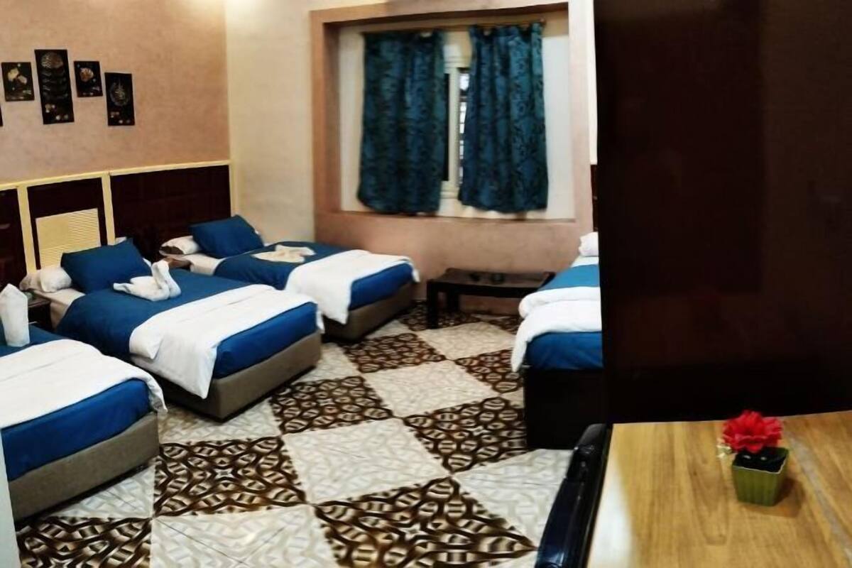 A hotel room with two beds, a window with curtains, a patterned floor, and a small table with a vase.