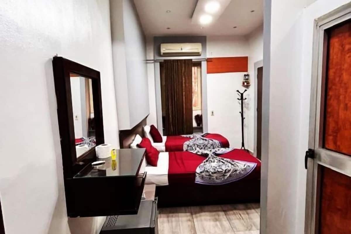 A narrow hotel room with a bed, a desk, a mirror, and an air conditioning unit.