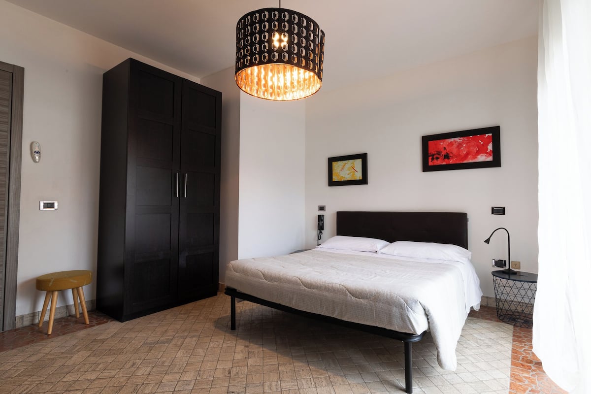 A bedroom with a bed, a wardrobe, a stool, a pendant light, a nightstand, and two framed pictures on the wall.