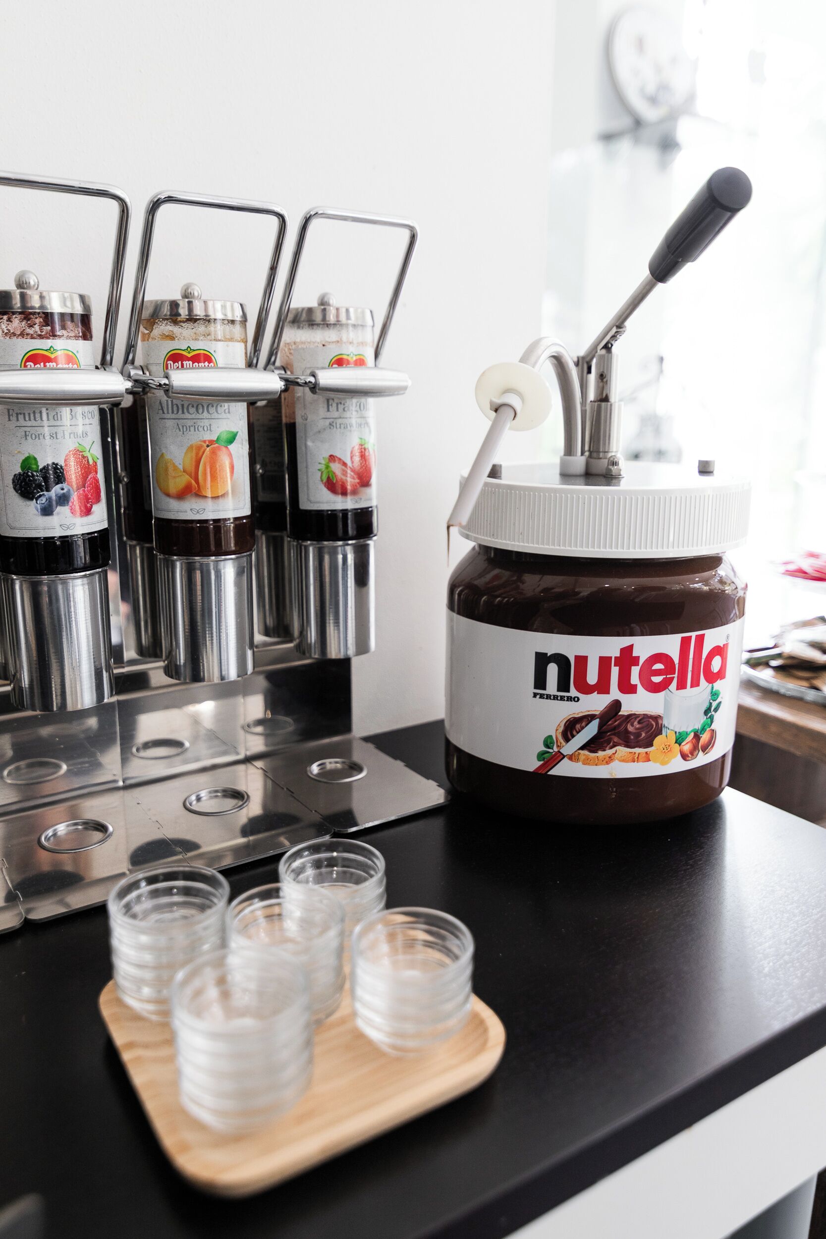 Un distributeur de Nutella avec plusieurs pots de différentes saveurs, une cuillère et de petits gobelets, le tout posé sur un comptoir noir.