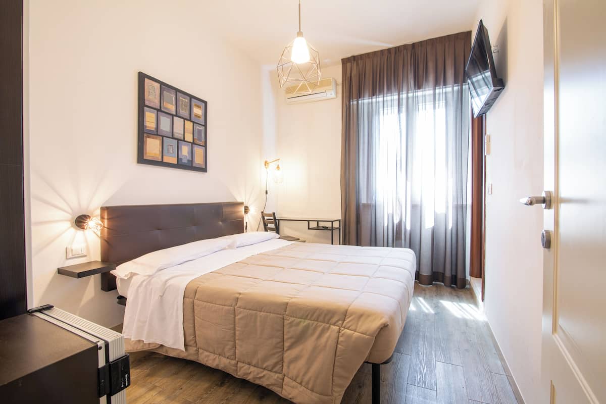 A hotel room with a bed, bedside table, lamp, artwork, and a TV mounted on the wall.