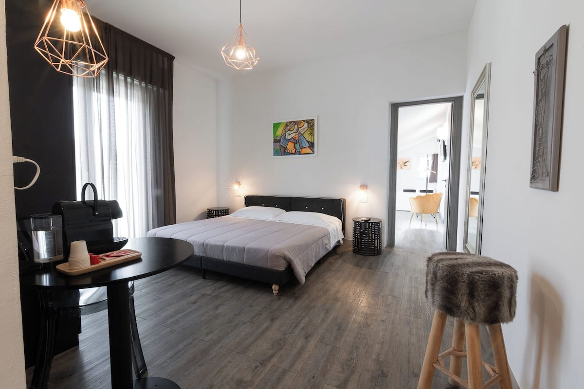 A modern hotel room with a large bed, a desk with a coffee maker, a chair, and a small stool.