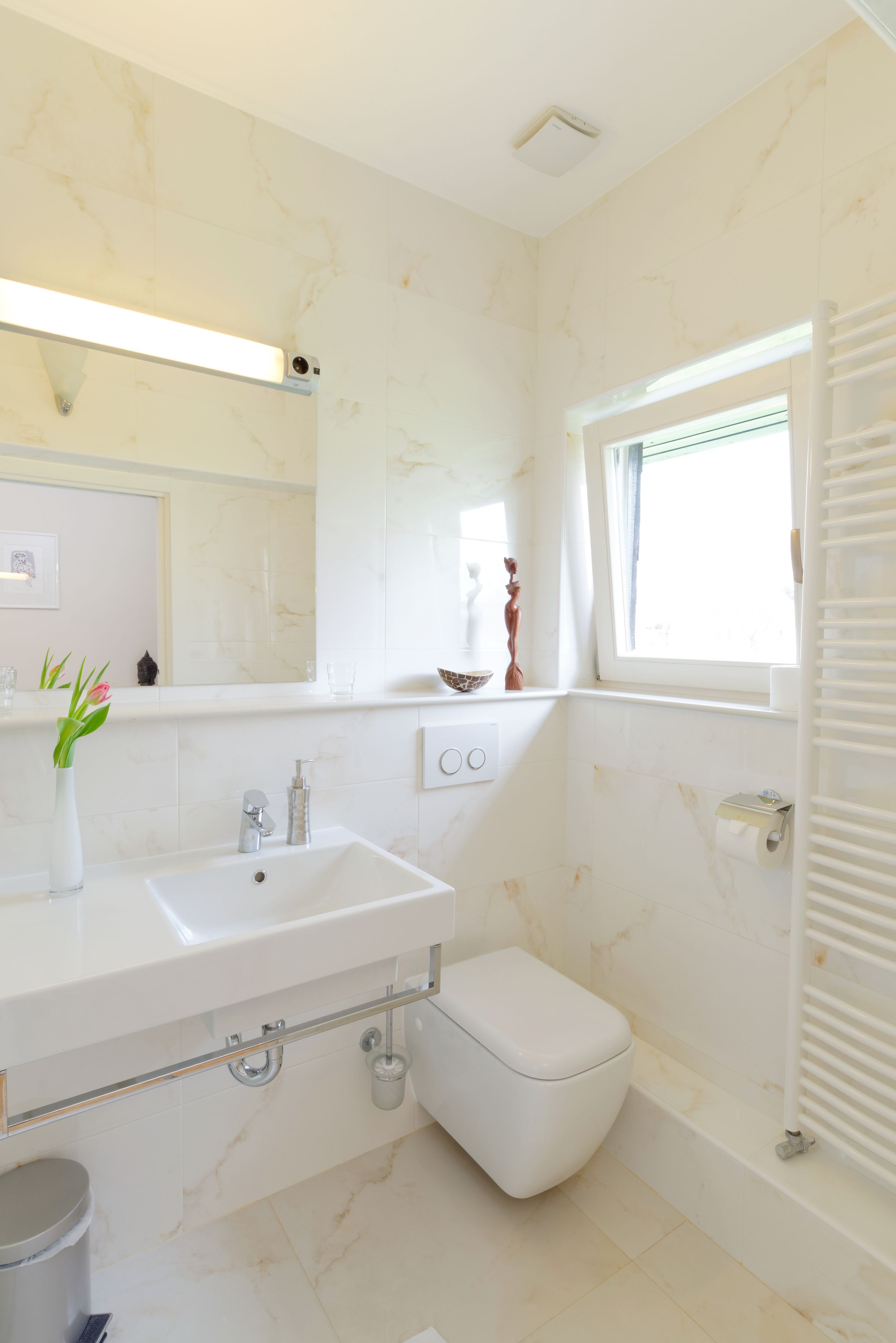 Une salle de bain moderne avec un lavabo blanc, un miroir et des toilettes. On y trouve une fenêtre avec des stores, un porte-serviettes et une petite étagère ornée d’objets décoratifs.
