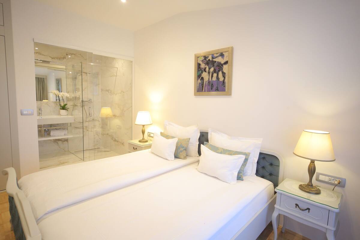A hotel room with a large bed, a glass shower enclosure, a bedside table with a lamp, and a painting on the wall.