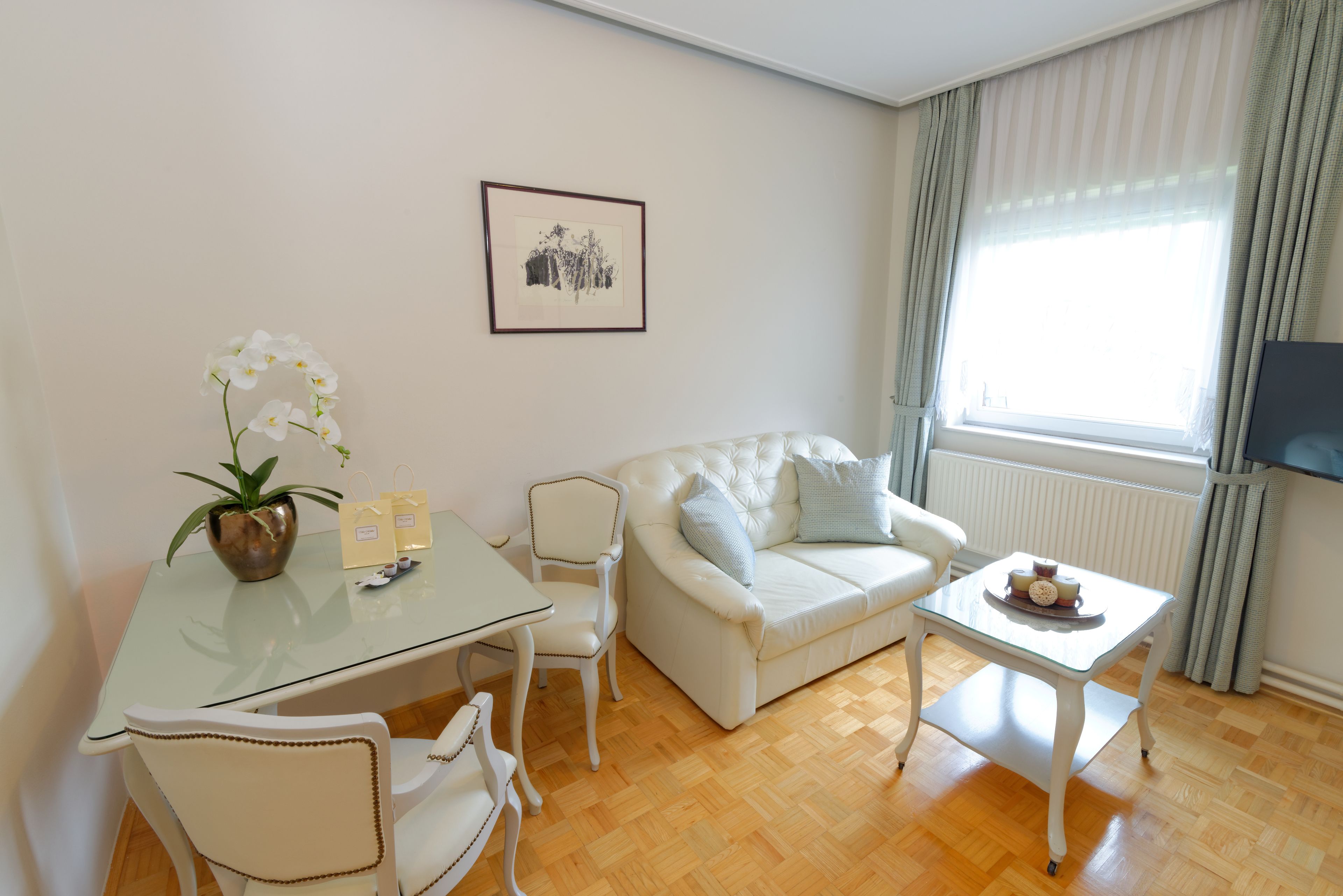 A living room with a white sofa, a glass coffee table, a white chair, a small table with a potted orchid, and a framed picture on the wall.