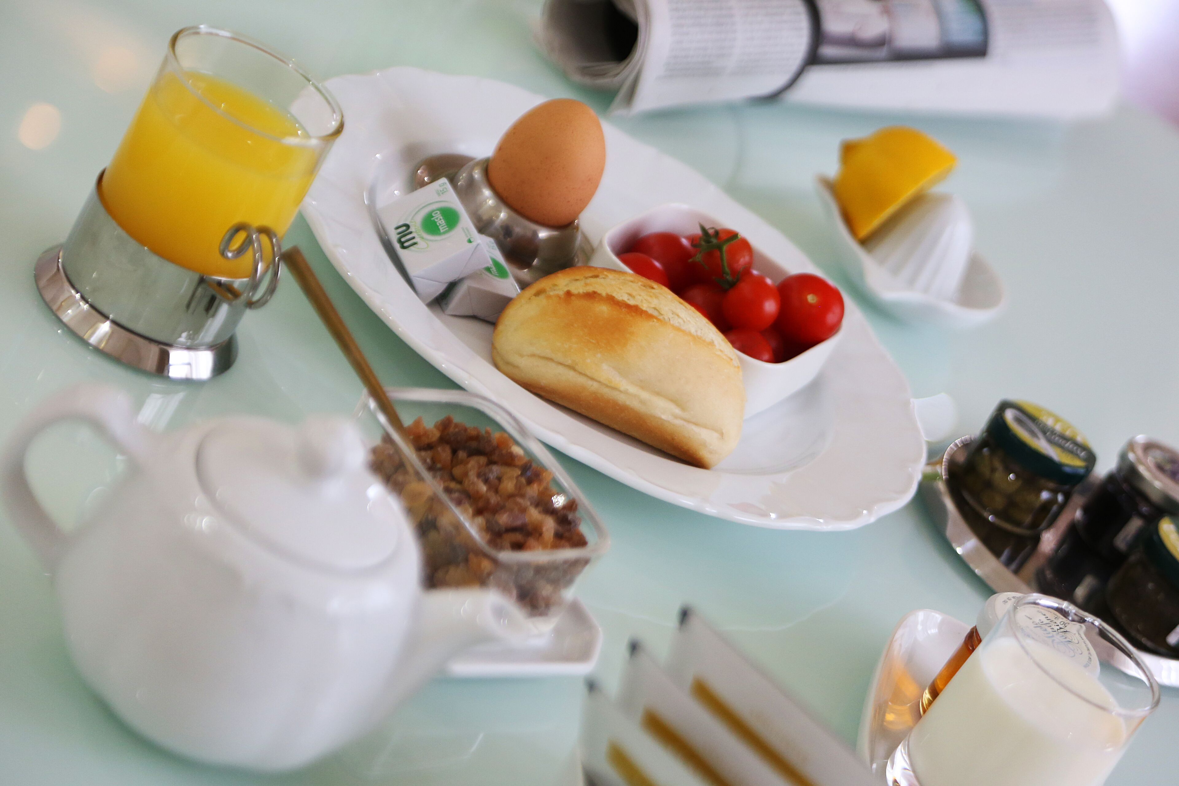 A breakfast setting with a teapot, a glass of orange juice, a plate with eggs, tomatoes, and bread, and condiments on a table.